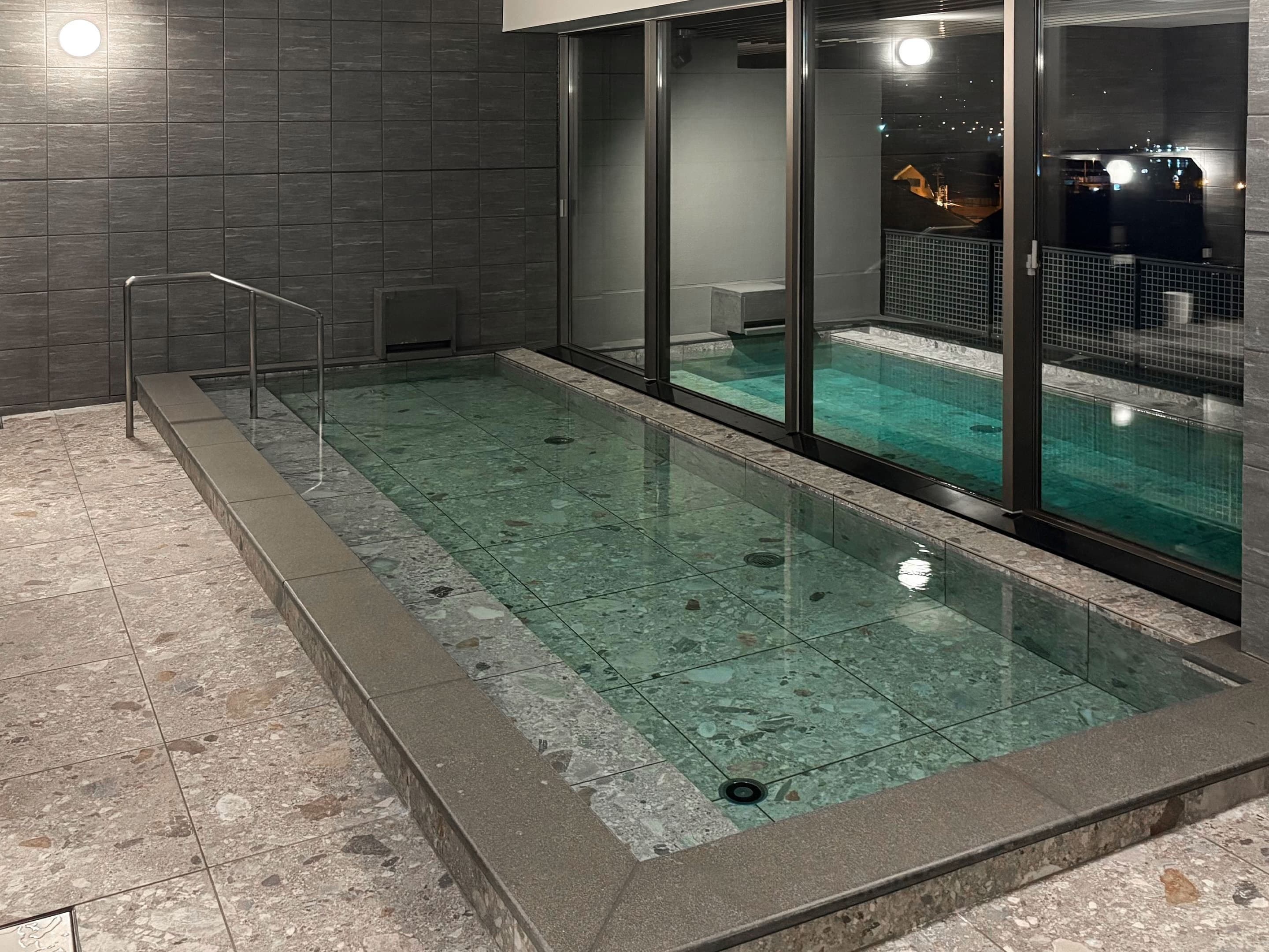 A swimming pool with clear water, surrounded by a stone-tiled deck and glass walls.