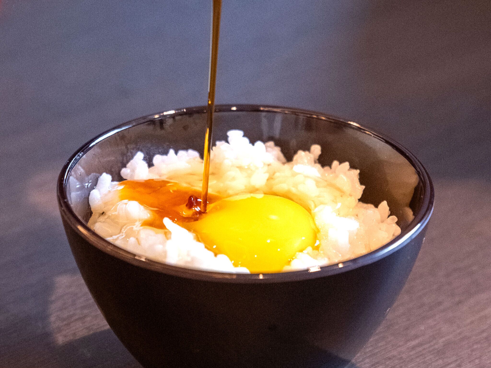 A bowl containing rice and a raw egg with a sauce being poured over it.
