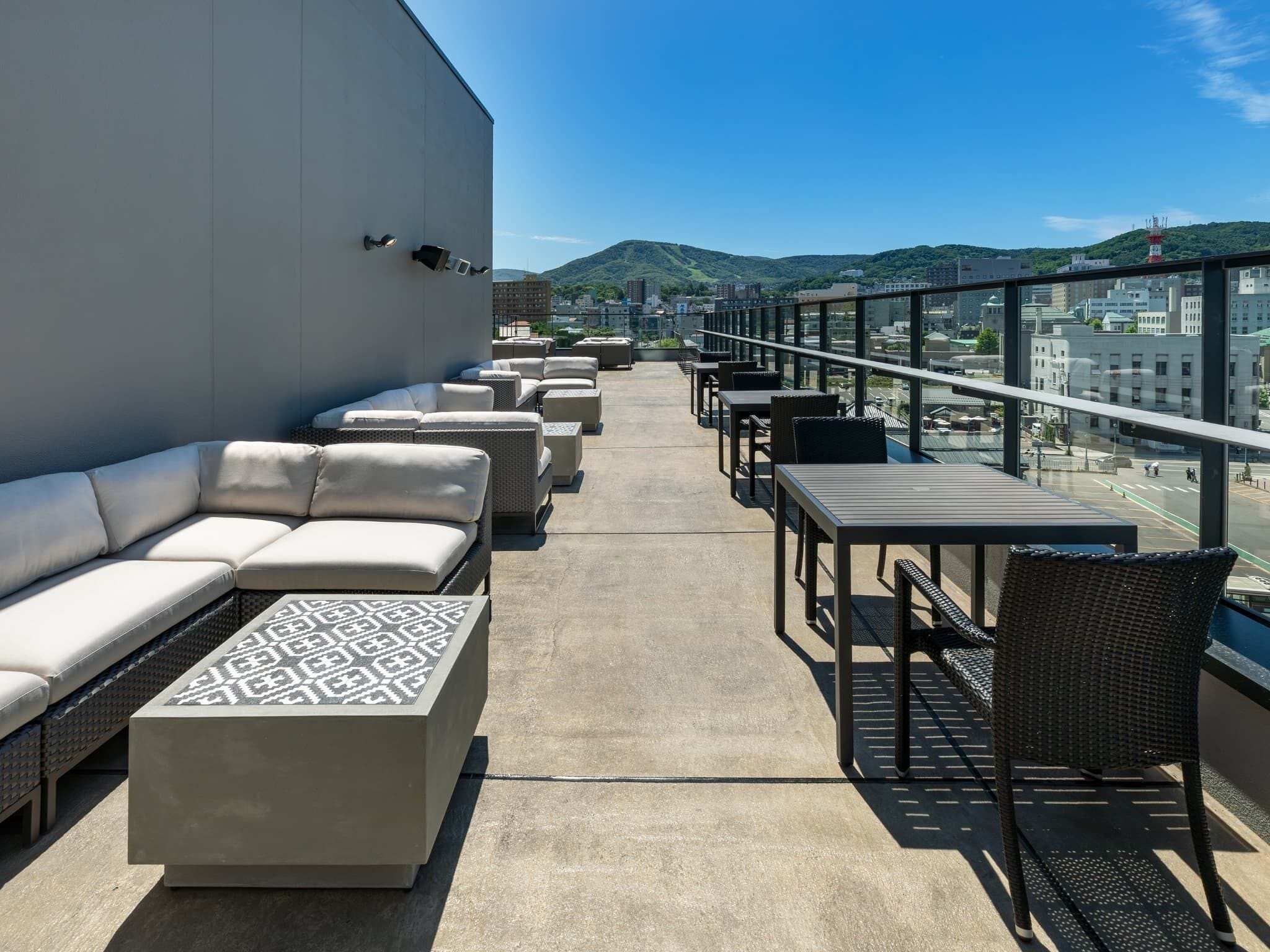 Una terraza en la azotea con sofá, sillas y mesas, que ofrece vistas a la ciudad y las colinas.