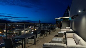 Een terras op het dak met buitenmeubilair, waaronder banken en tafels, met uitzicht op een stadsgezicht in de schemering.