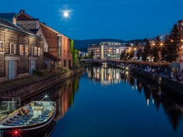 Un río tranquilo con un bote amarrado a la izquierda, un puente en el centro y edificios a ambos lados bajo un cielo nocturno.