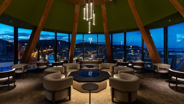 Een moderne lounge met een centrale tafel, omringd door banken, grote ramen met uitzicht over de stad, en een houten plafondconstructie.