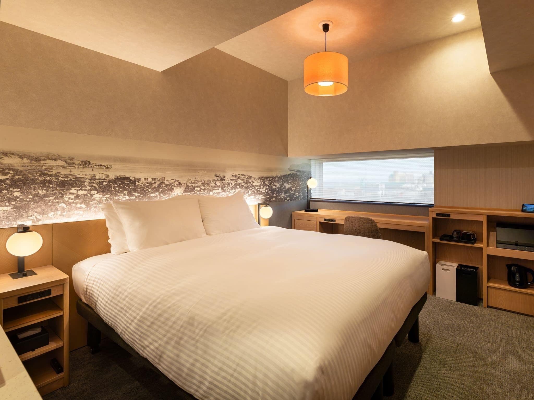 Ein modernes Hotelzimmer mit einem großen Bett, einem Schreibtisch, einem Stuhl, einer Lampe, einer kleinen Ablage mit einer Kaffeemaschine und einem Wandbild mit Stadtansicht.