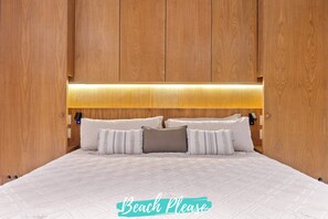 Ein ordentlich bezogenes Bett mit weißer Steppdecke und mehreren Kissen, ein Holz-Kopfteil mit einer Beleuchtung und ein Schild mit der Aufschrift 'Beach Please' am Fußende.