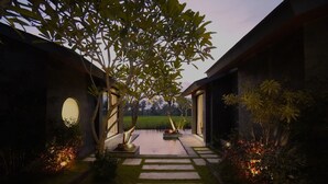 Un chemin menant à un bâtiment doté d’une fenêtre circulaire, entouré d’une végétation luxuriante et d’un petit étang.