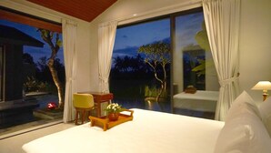 Une chambre avec un grand lit, une table de chevet avec un plateau, une chaise et une vue sur une piscine et des arbres à l’extérieur.