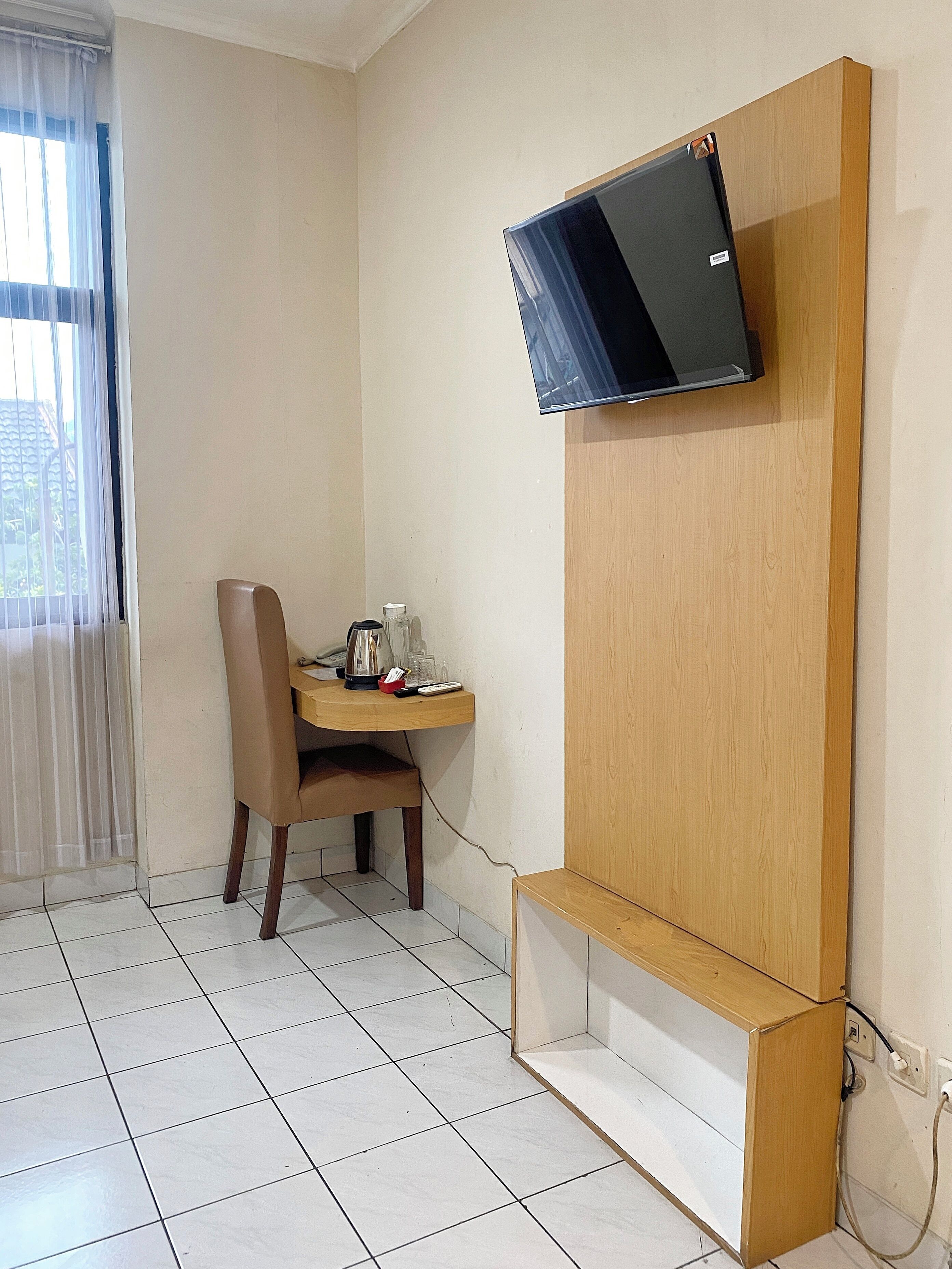 A small room with a wooden TV stand, a chair, a small table with a kettle, and a window with sheer curtains.