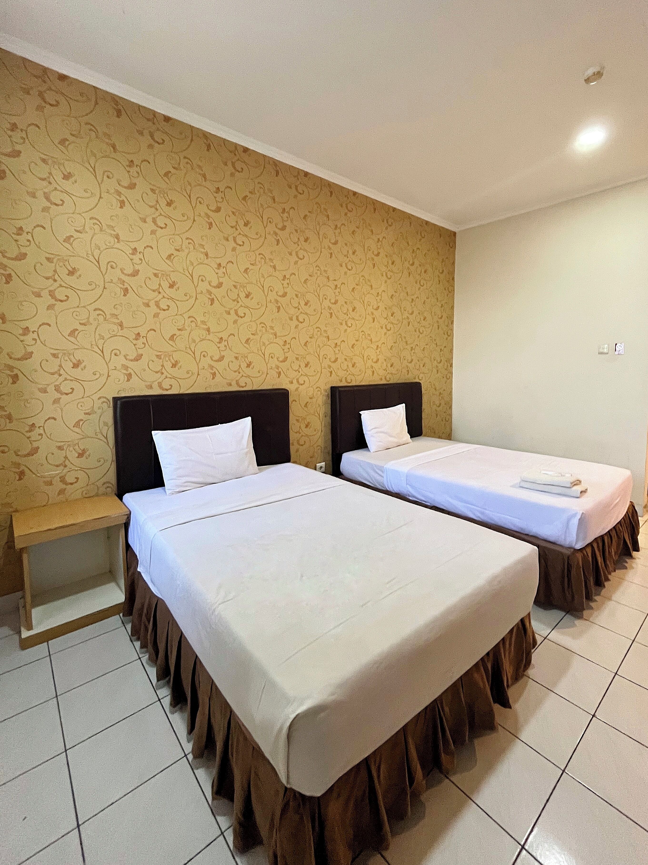 Habitación de hotel con dos camas, una mesita de noche pequeña y papel tapiz estampado.