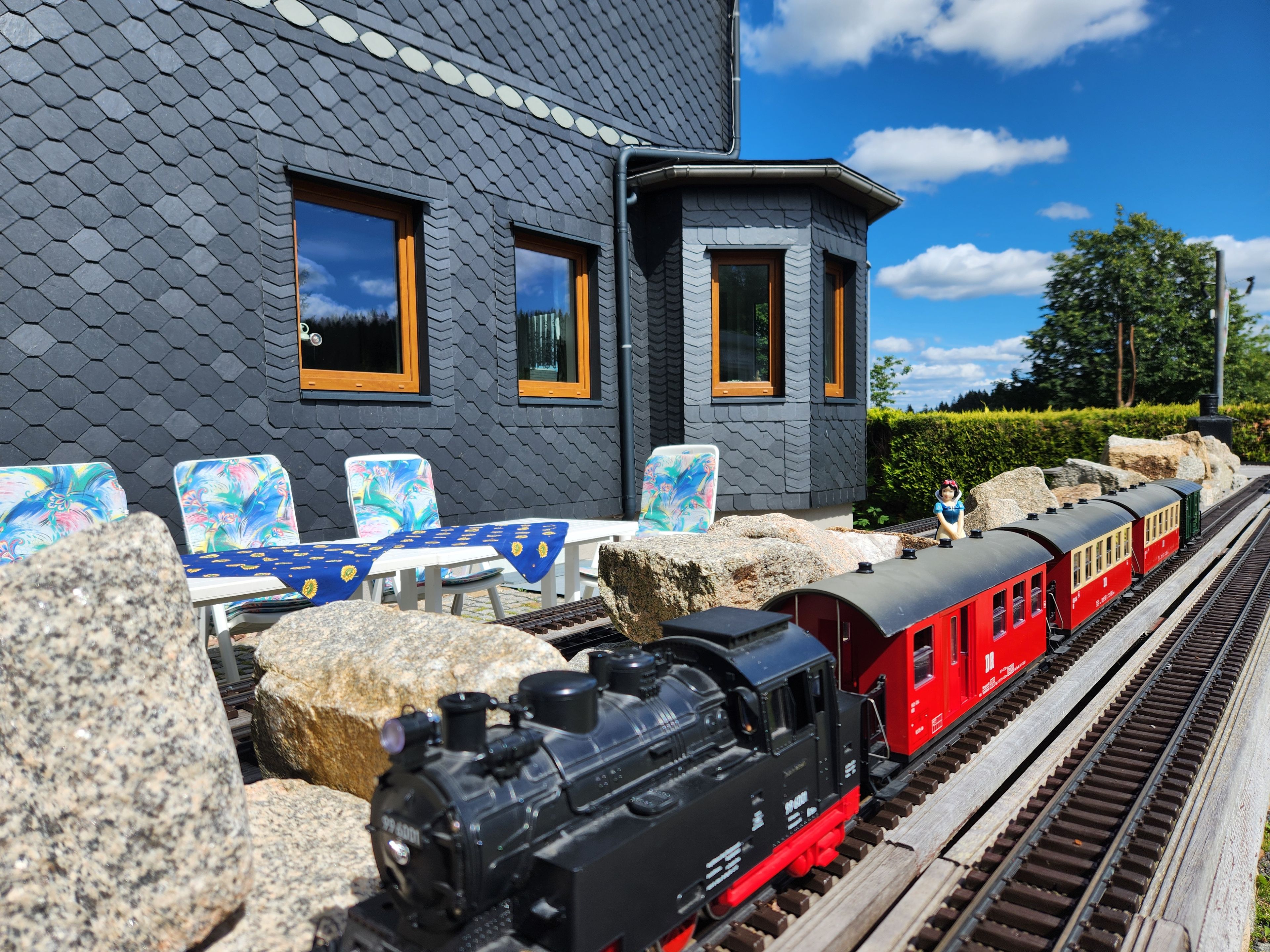 Ein Modelleisenbahn-Set mit einer schwarzen Lokomotive und roten Personenwagen auf Schienen, ein weißer Tisch mit blau-orangen Stühlen mit Muster und ein graues Gebäude mit Fenstern.