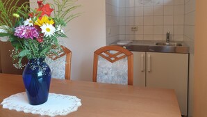 A dining table with a blue vase of flowers, a wooden chair, and a kitchen with white tiles and a sink. - Siegmundsburger Haus Werraquelle here Apartment "Traumwelt" family- / pet-friendly (Siegmundsburg)