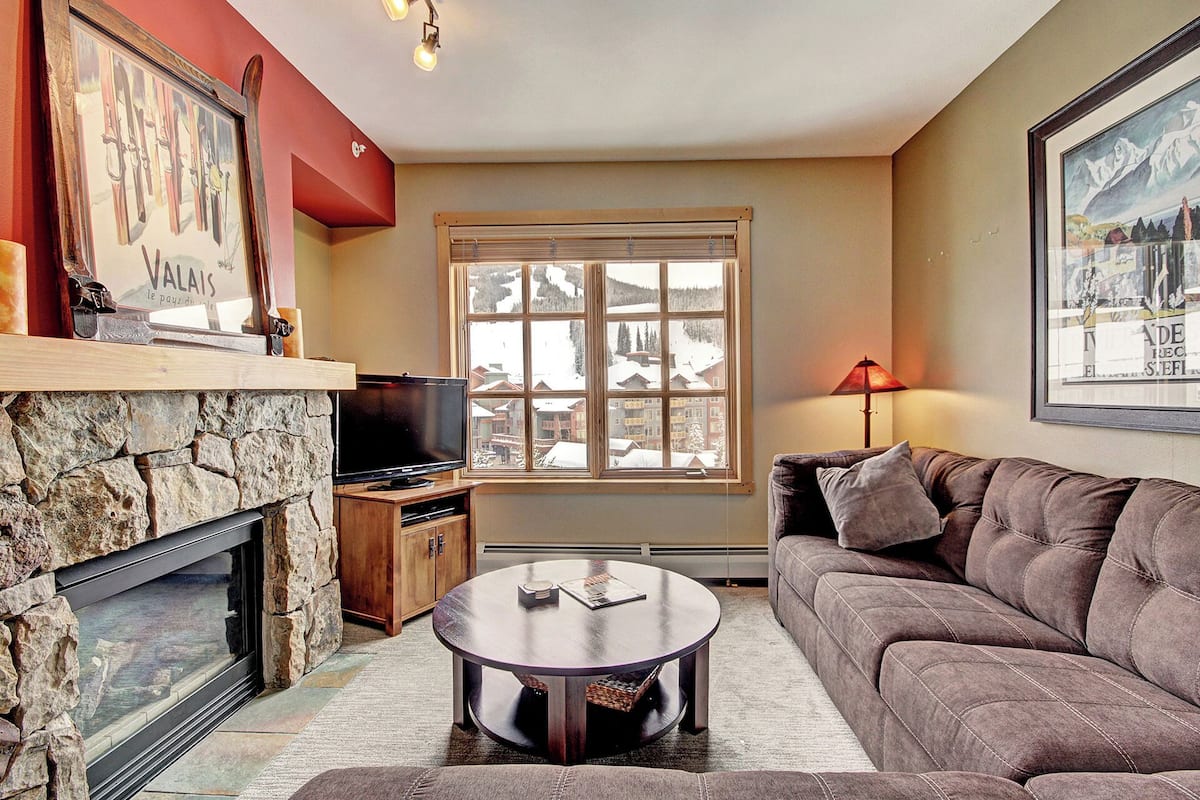 A cozy living room with a stone fireplace, a flat-screen TV, a comfortable sofa, and a large window offering a view of a snowy village.