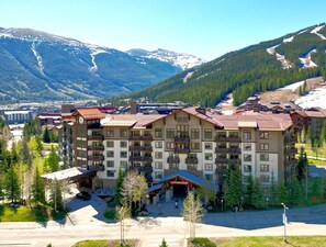 Un hotel de varios pisos con una entrada central, rodeado de montañas nevadas y un bosque denso.