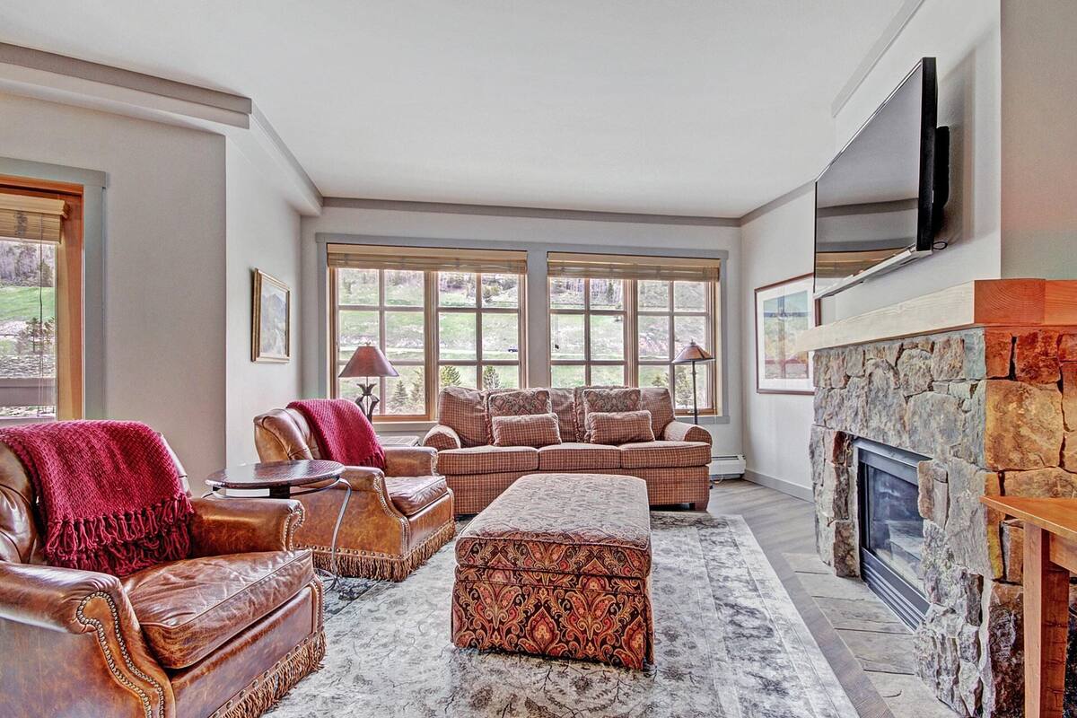 A spacious living room with leather sofas, a stone fireplace, and large windows offering a view of the outdoors.