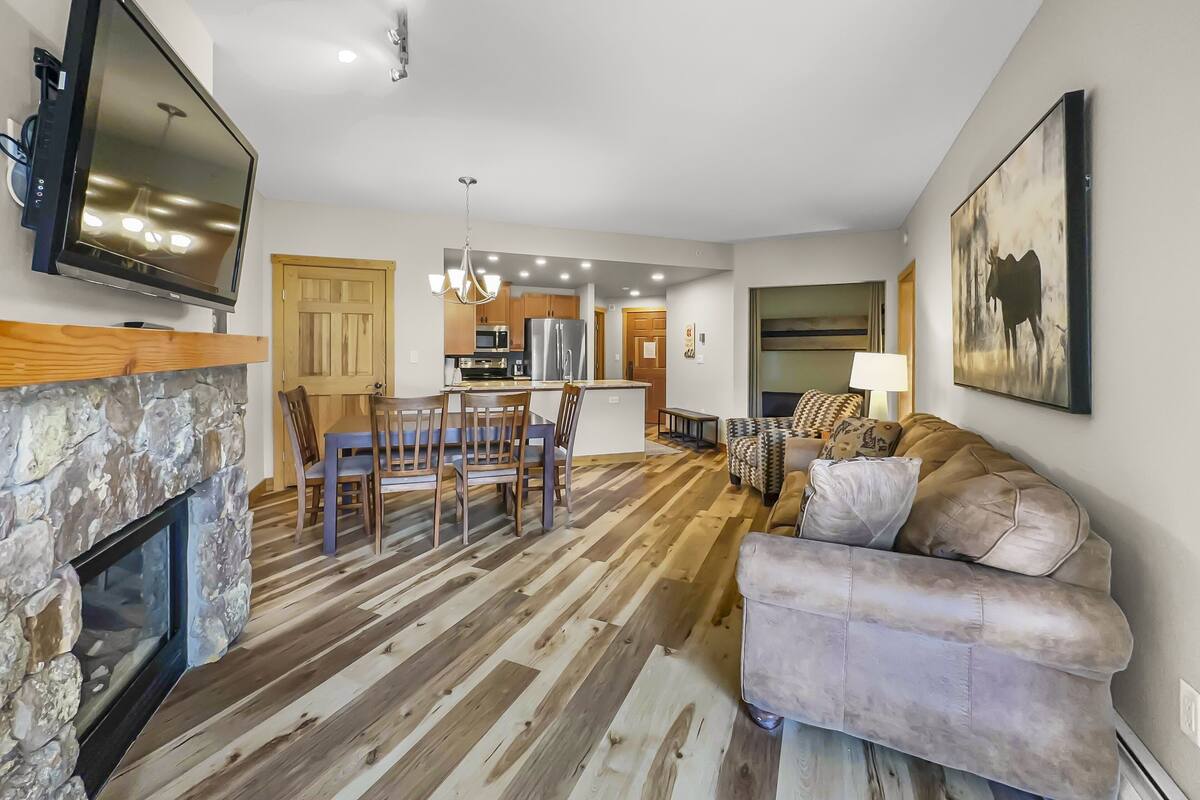 A modern living room with a stone fireplace, a kitchen with wooden flooring, a dining area, and a large sofa.