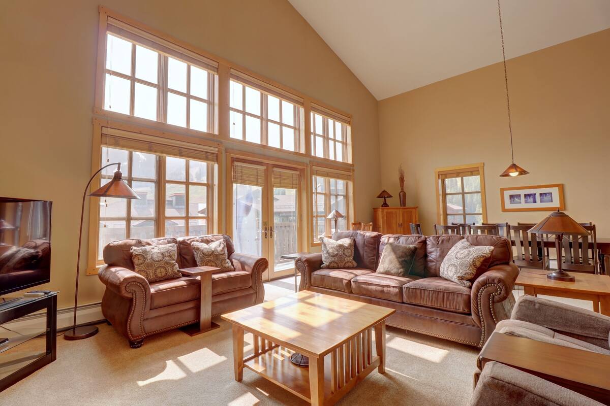 A spacious living room with large windows, leather sofas, a wooden coffee table, and a television.
