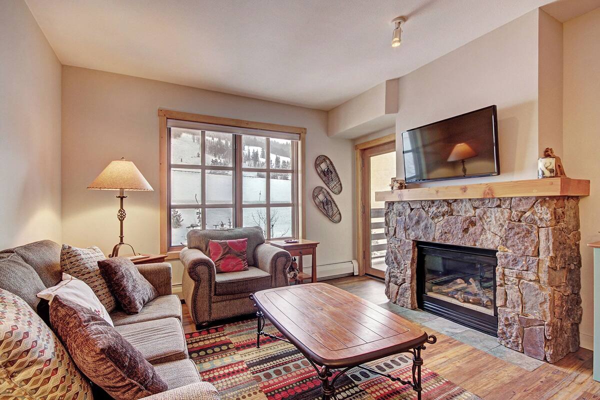 A cozy living room with a stone fireplace, a large window with a view of a snowy landscape, a comfortable sofa, and a flat-screen TV mounted above the fireplace.