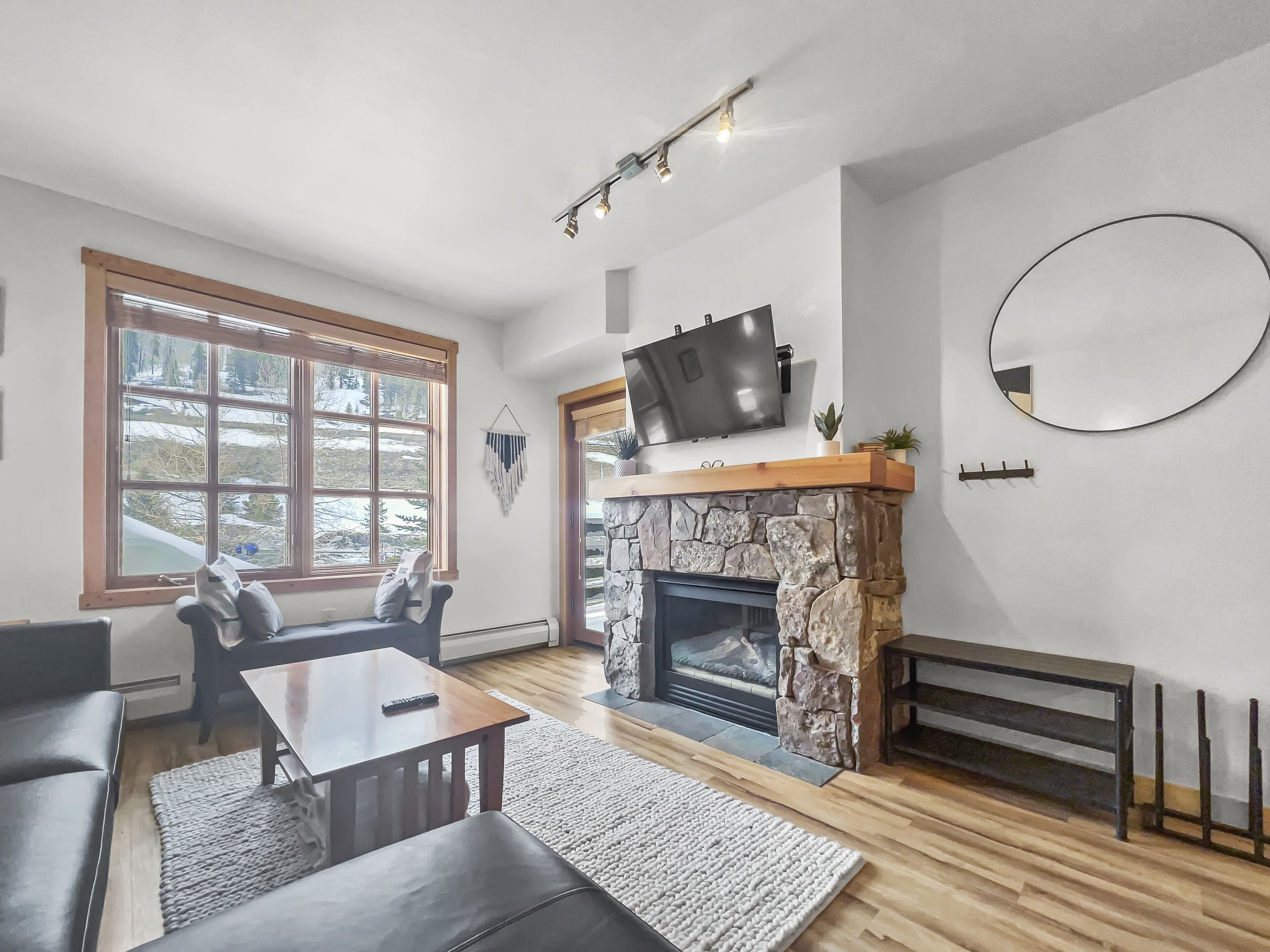 Een moderne woonkamer met een stenen open haard, een flatscreen televisie erboven en een ronde spiegel aan de muur, met een groot raam en uitzicht op bomen.