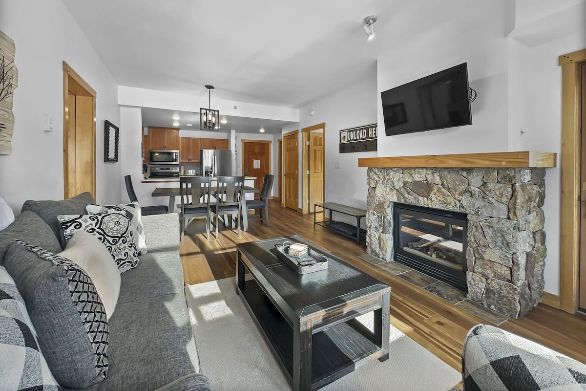 A modern living room with a fireplace, a dining area, and a kitchen in the background.