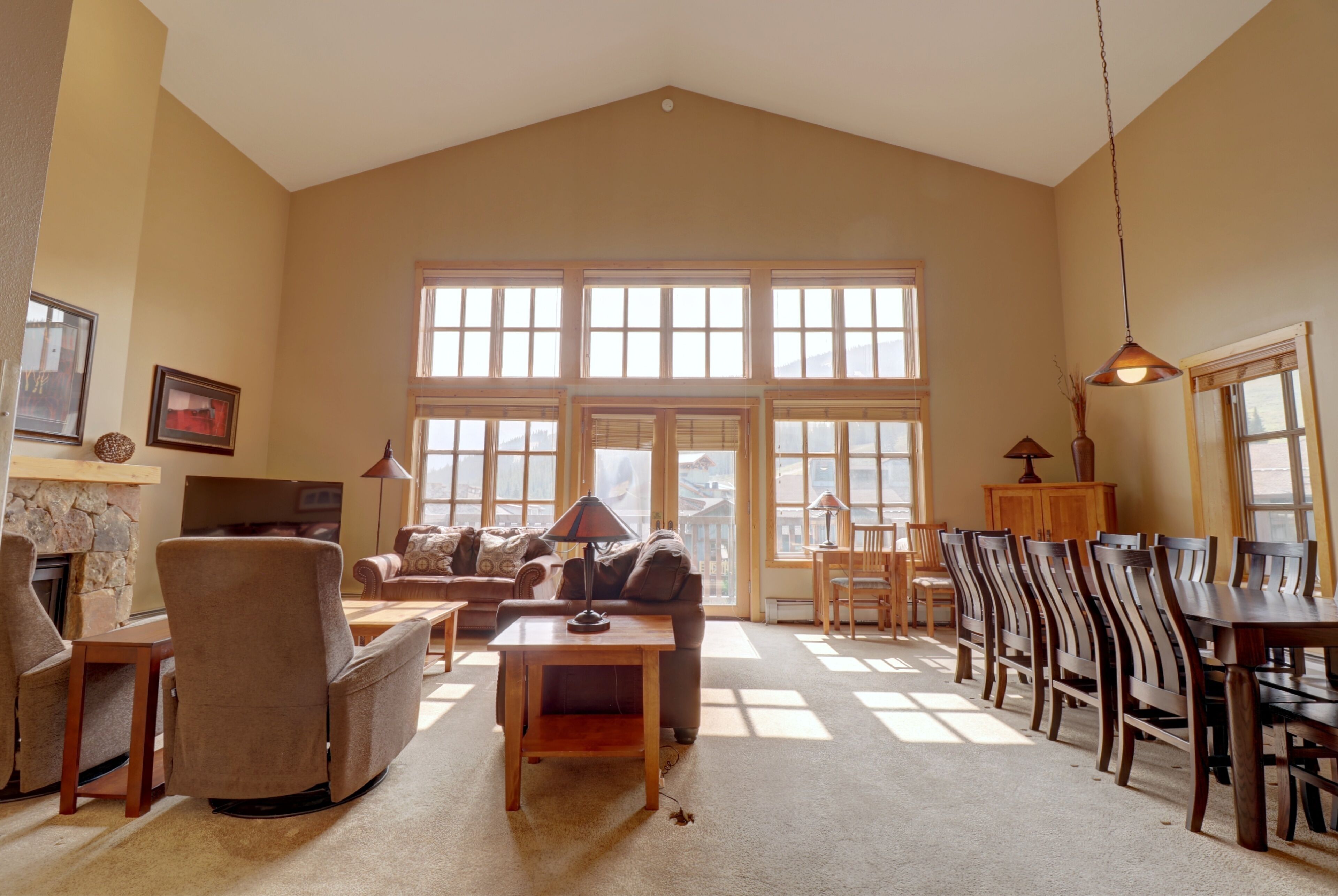A spacious living room with a high ceiling, large windows, a fireplace, and a dining area with wooden furniture.