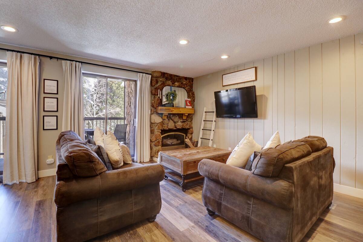 A living room with two brown leather sofas, a wooden coffee table, a stone fireplace, a mounted TV, and a window with curtains.
