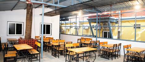 A spacious indoor area with multiple wooden tables and chairs arranged in rows, large windows allowing natural light, and a high ceiling with metal beams.