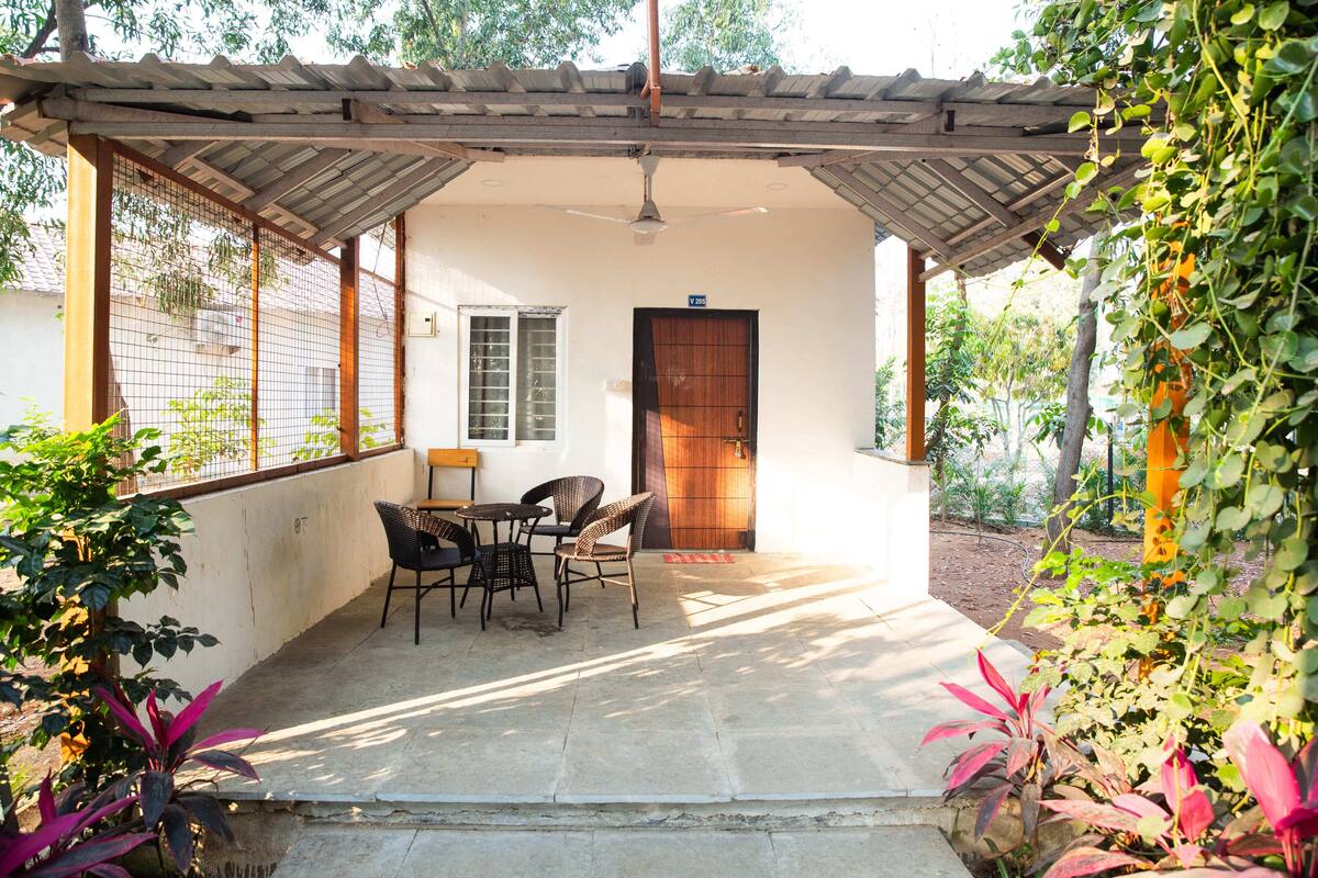 A covered patio with a table and chairs, a wooden door, and a view of the surrounding greenery.