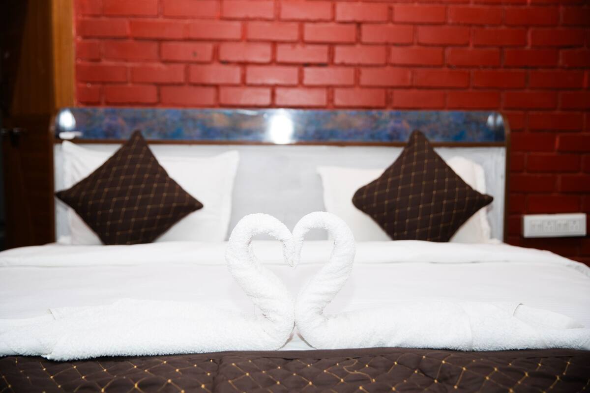 A bed with white linens, a heart-shaped towel arrangement, and a red brick wall in the background.