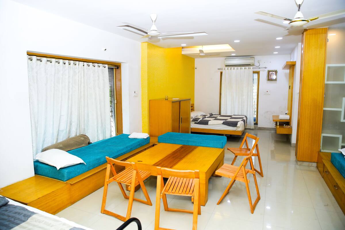 A compact hotel room with a sofa bed, a small table with chairs, a ceiling fan, and an air conditioning unit.