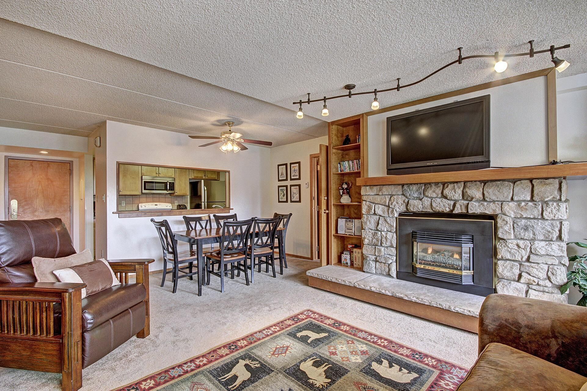 A living room with a stone fireplace, a dining area with a kitchen in the background, and a ceiling fan.