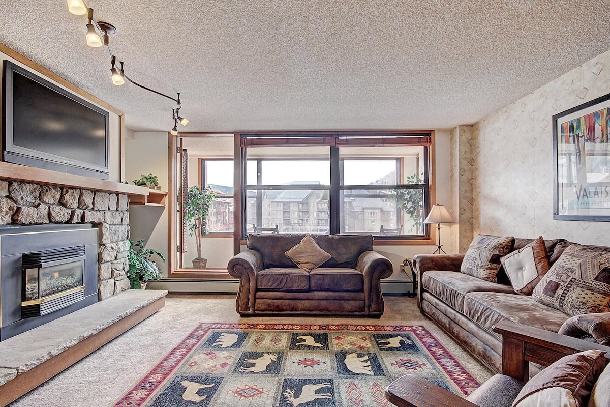 Ein gemütliches Wohnzimmer mit Kamin, einer braunen Ledersofa, einem gemusterten Teppich und einem großen Fenster mit Blick auf Gebäude.