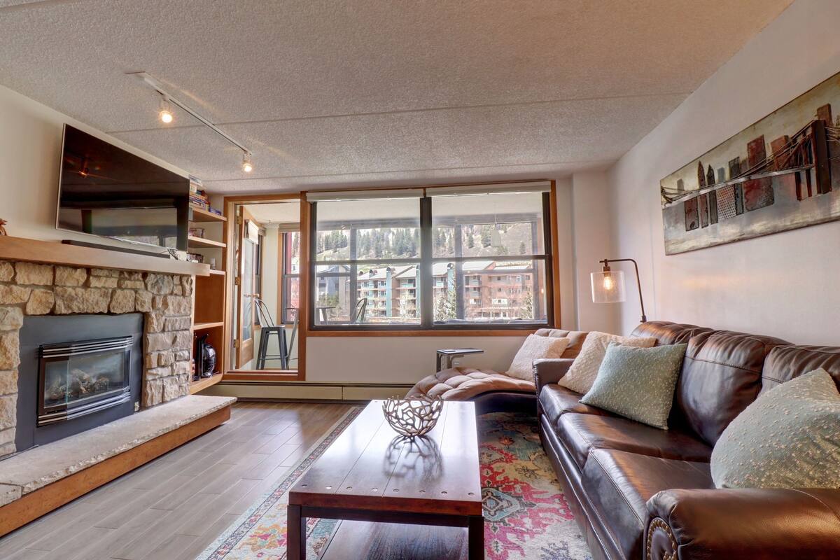 Ein Wohnzimmer mit Kamin aus Stein, einer Ledersofa, einem Couchtisch und einem großen Fenster mit Blick auf Gebäude.