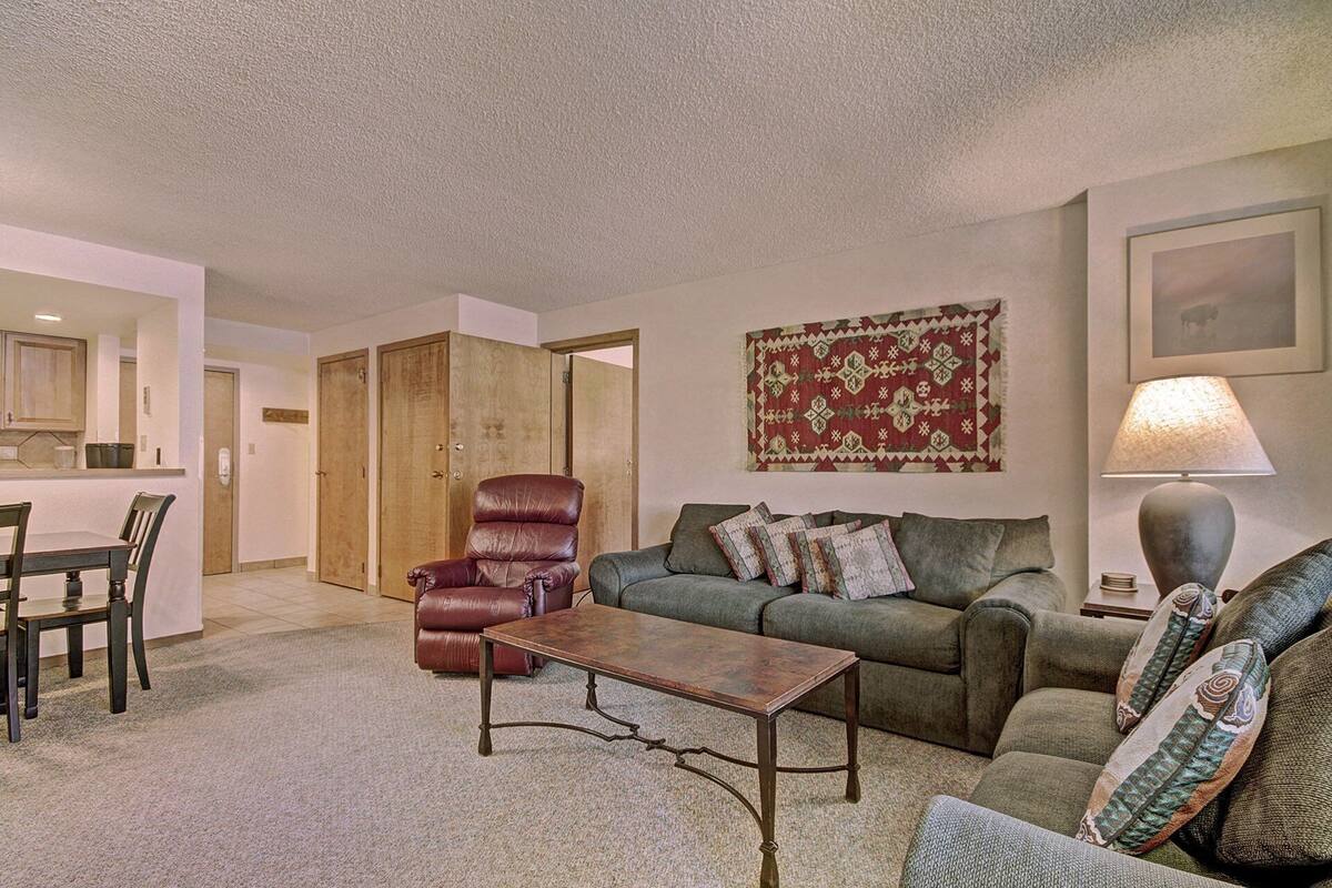 Ein Wohnzimmer mit einer Couch, einem Sessel und einem Couchtisch. Es gibt einen Essbereich mit Stühlen und einem Tisch. Im Hintergrund ist eine Küche zu sehen.
