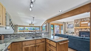 A spacious kitchen with wooden cabinets, a granite countertop, and a large island. There is a dining area with a blue sofa and a stone fireplace. - SkyRun Vacation Condos Copper Mountain Resort (Copper Mountain)