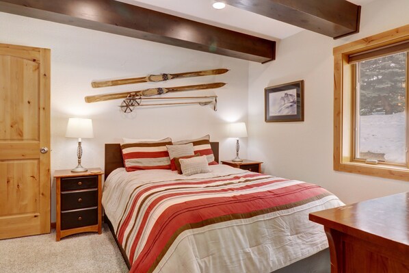 Une chambre à coucher avec un lit, une tête de lit en bois, des tables de chevet avec des lampes, une commode en bois, des skis accrochés au mur et une fenêtre donnant sur un paysage enneigé.