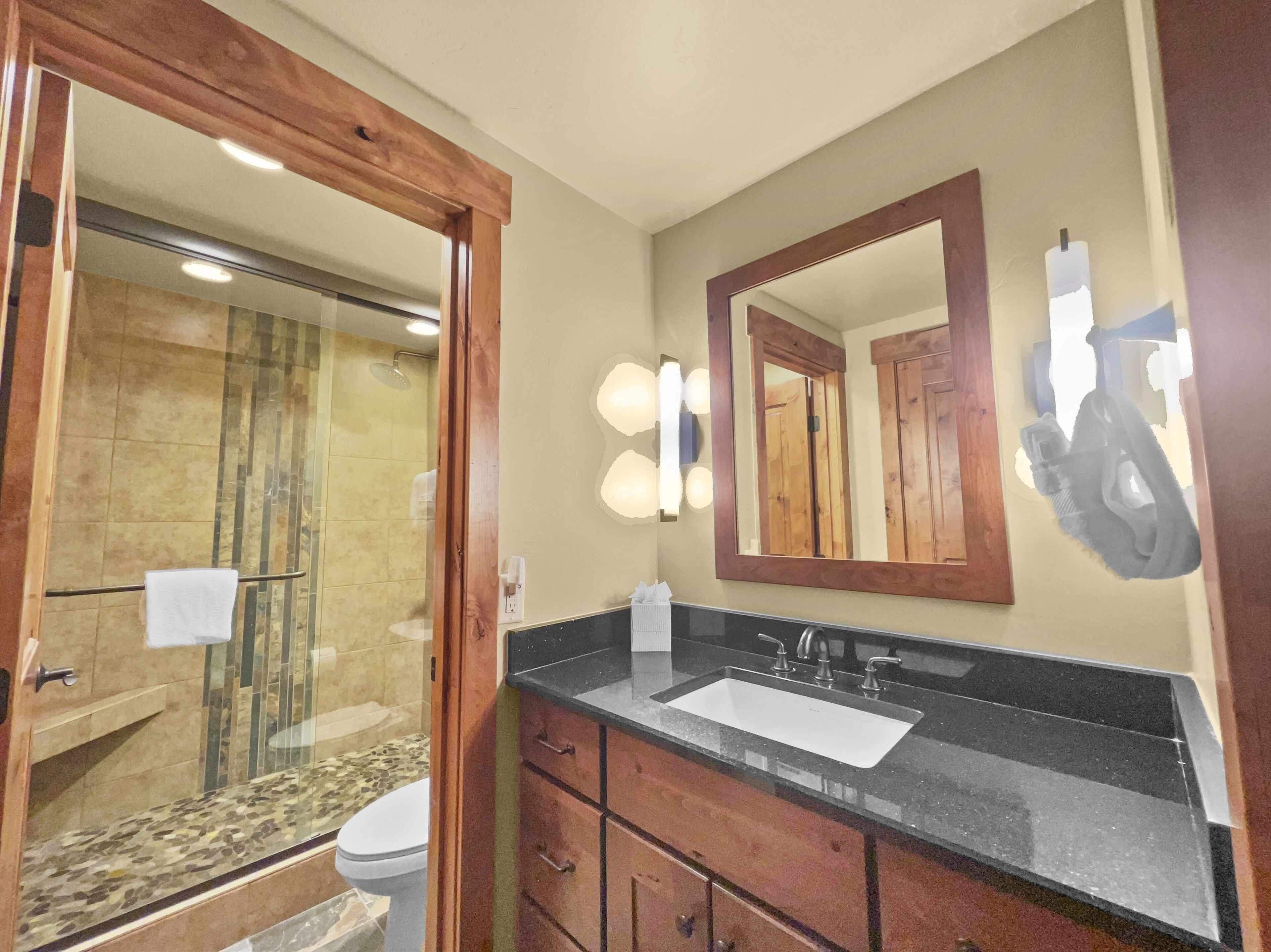A bathroom with a glass-enclosed shower, a sink with a black countertop, and a mirror with lights.