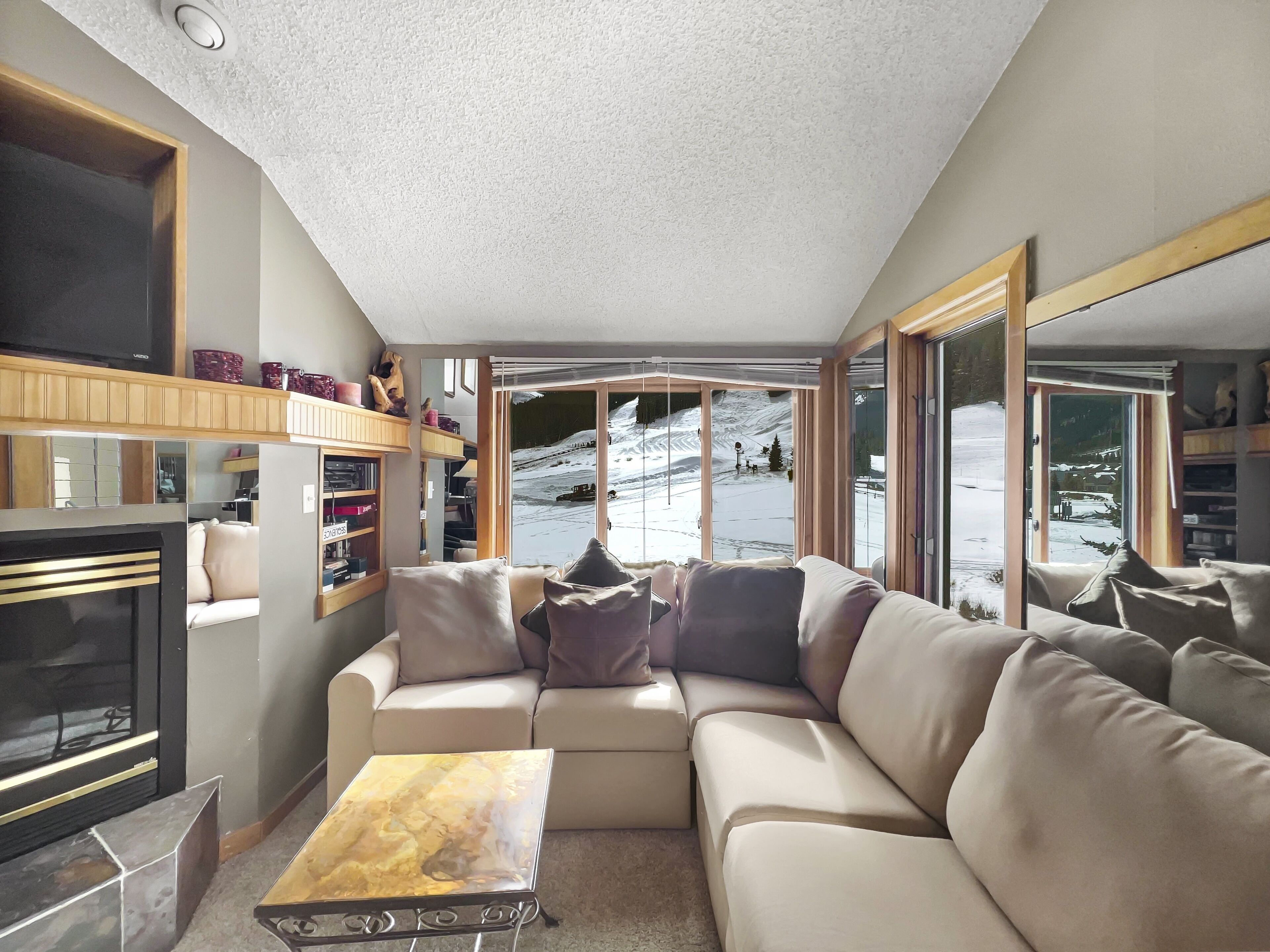A cozy living room with a sectional sofa, a fireplace, and a view of a snowy landscape through large windows.