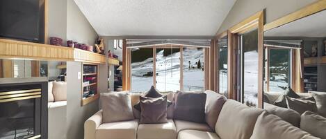 A cozy living room with a sectional sofa, a fireplace, and a view of a snowy landscape through large windows.