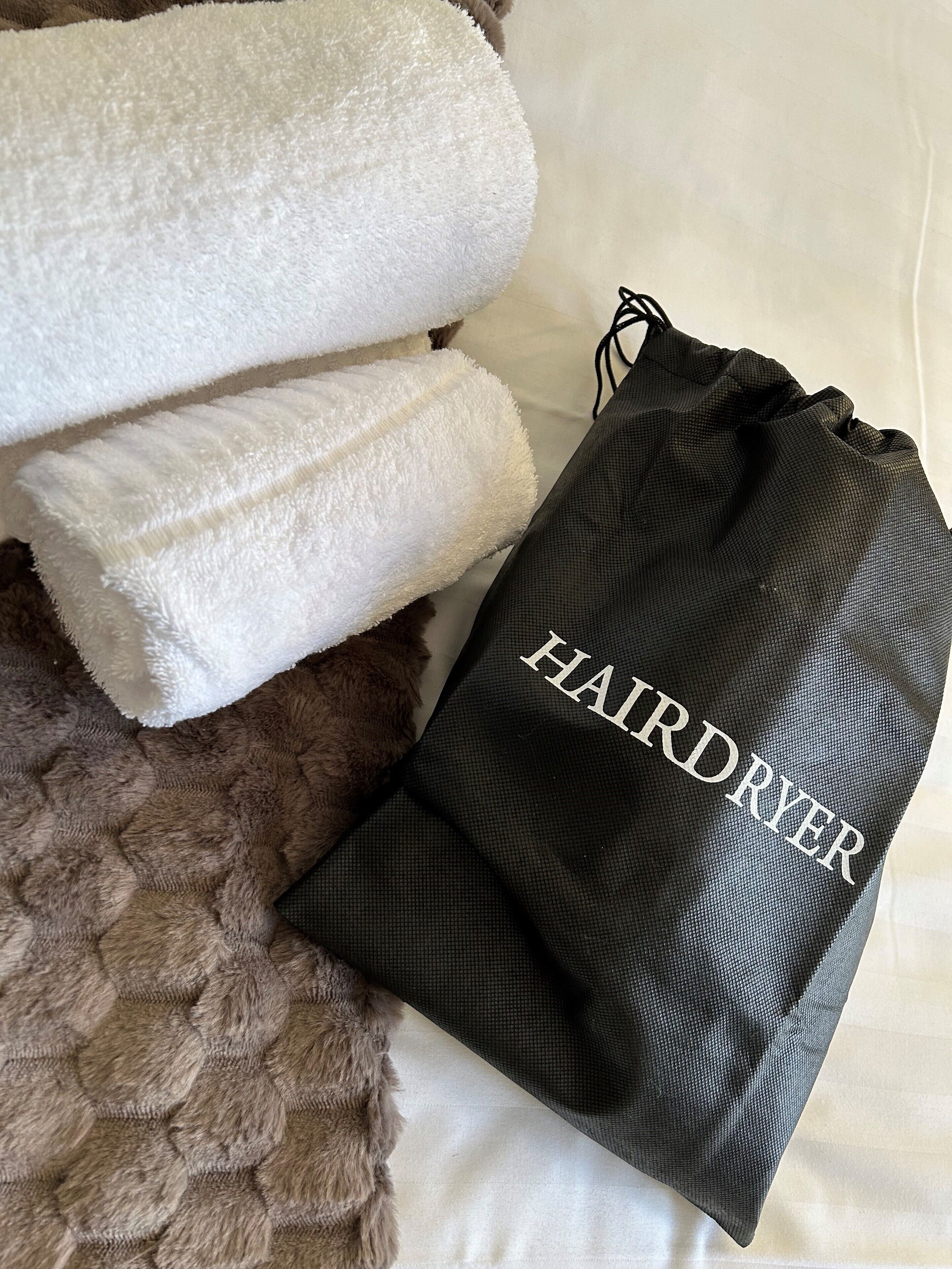 A bed with a white blanket and a black drawstring bag labeled "HAIRDRYER".