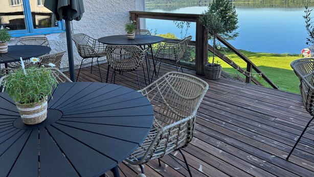 Une terrasse en bois avec des meubles d’extérieur, une vue sur un lac et un bâtiment à la façade blanche.