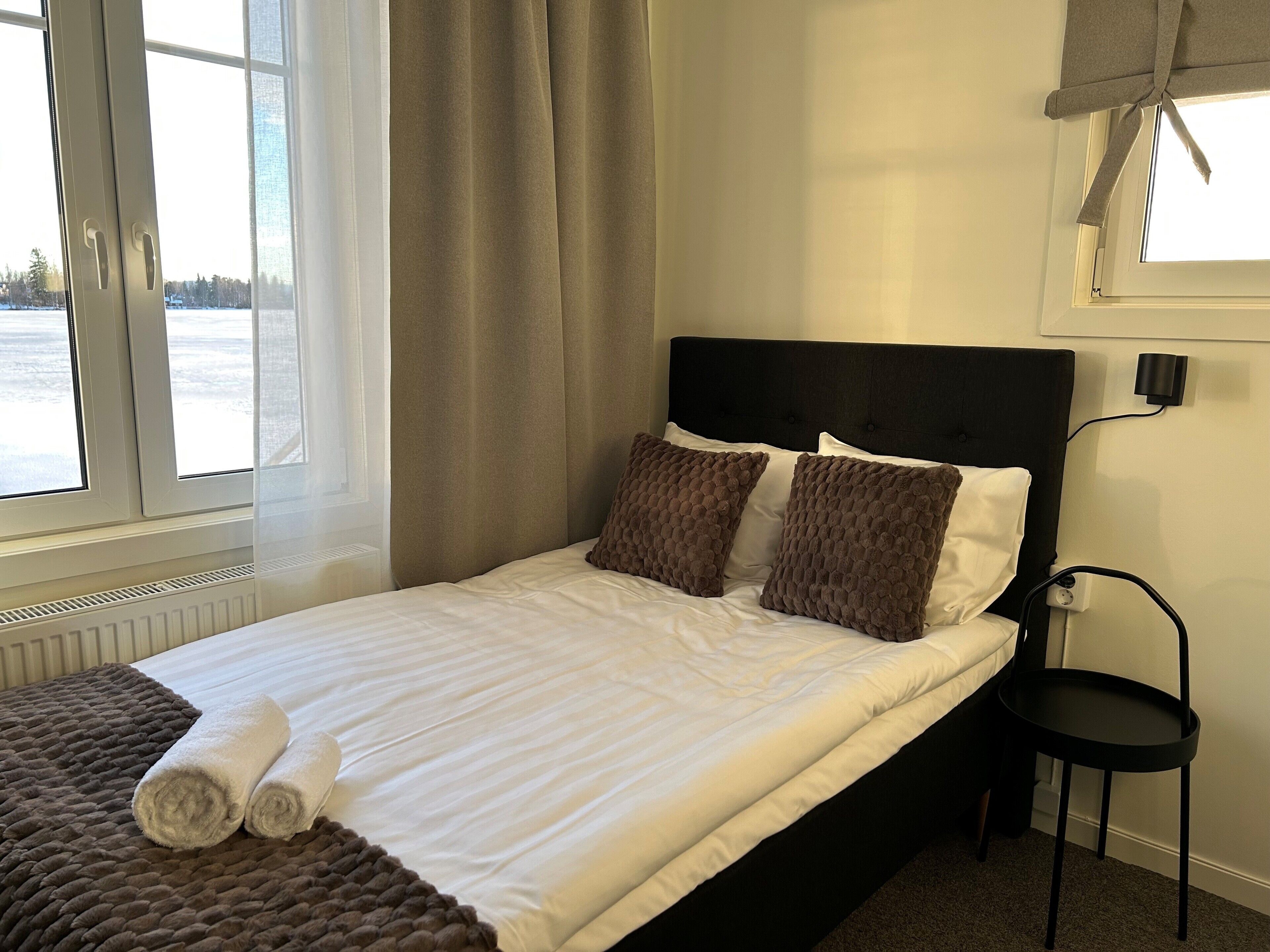 En pænt redt seng med hvide sengetæpper og to brune puder, en sort sengegavl og et lille natbord med en lampe.