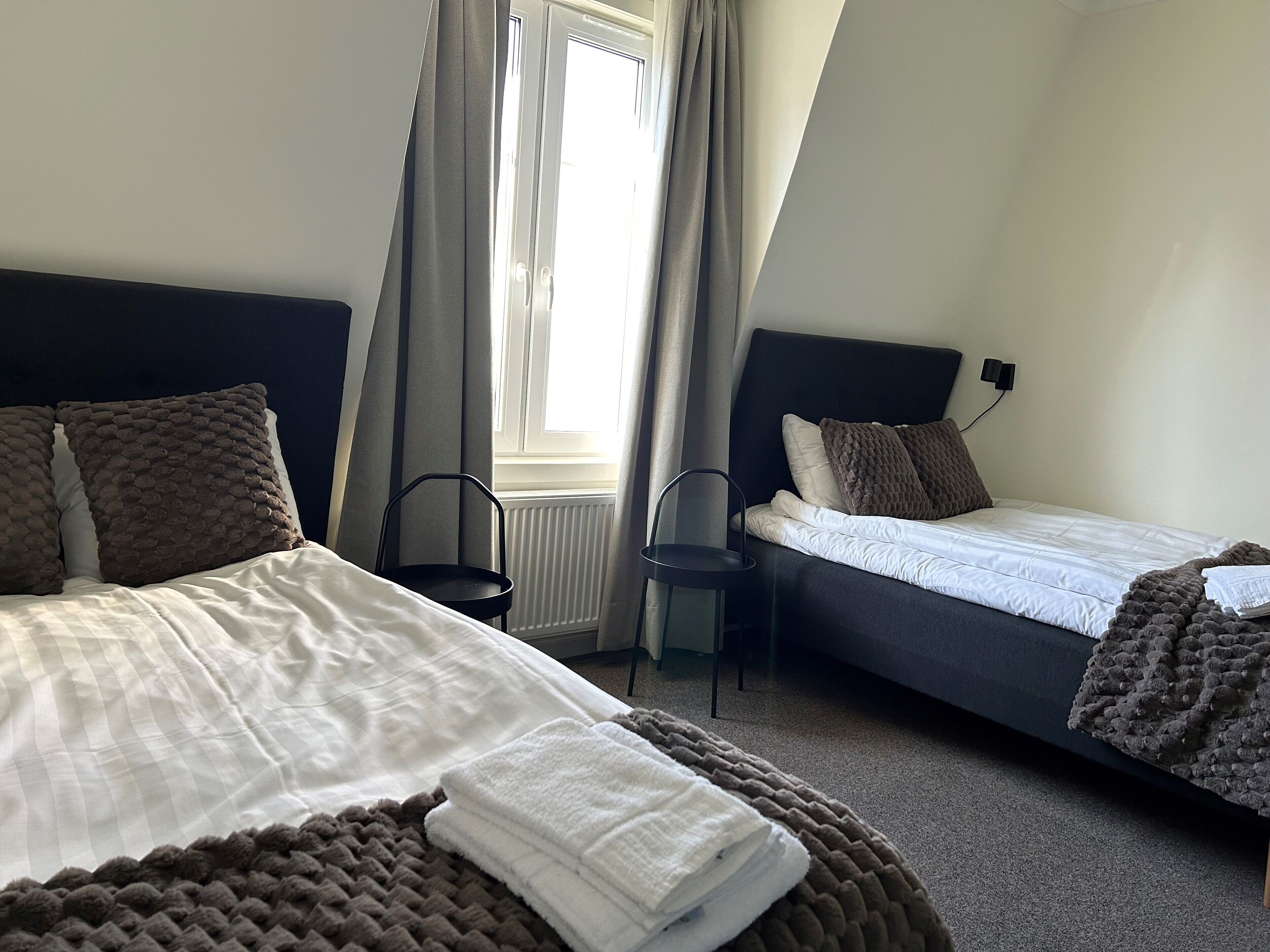 A hotel room with two beds, a window with curtains, a radiator, and a chair.