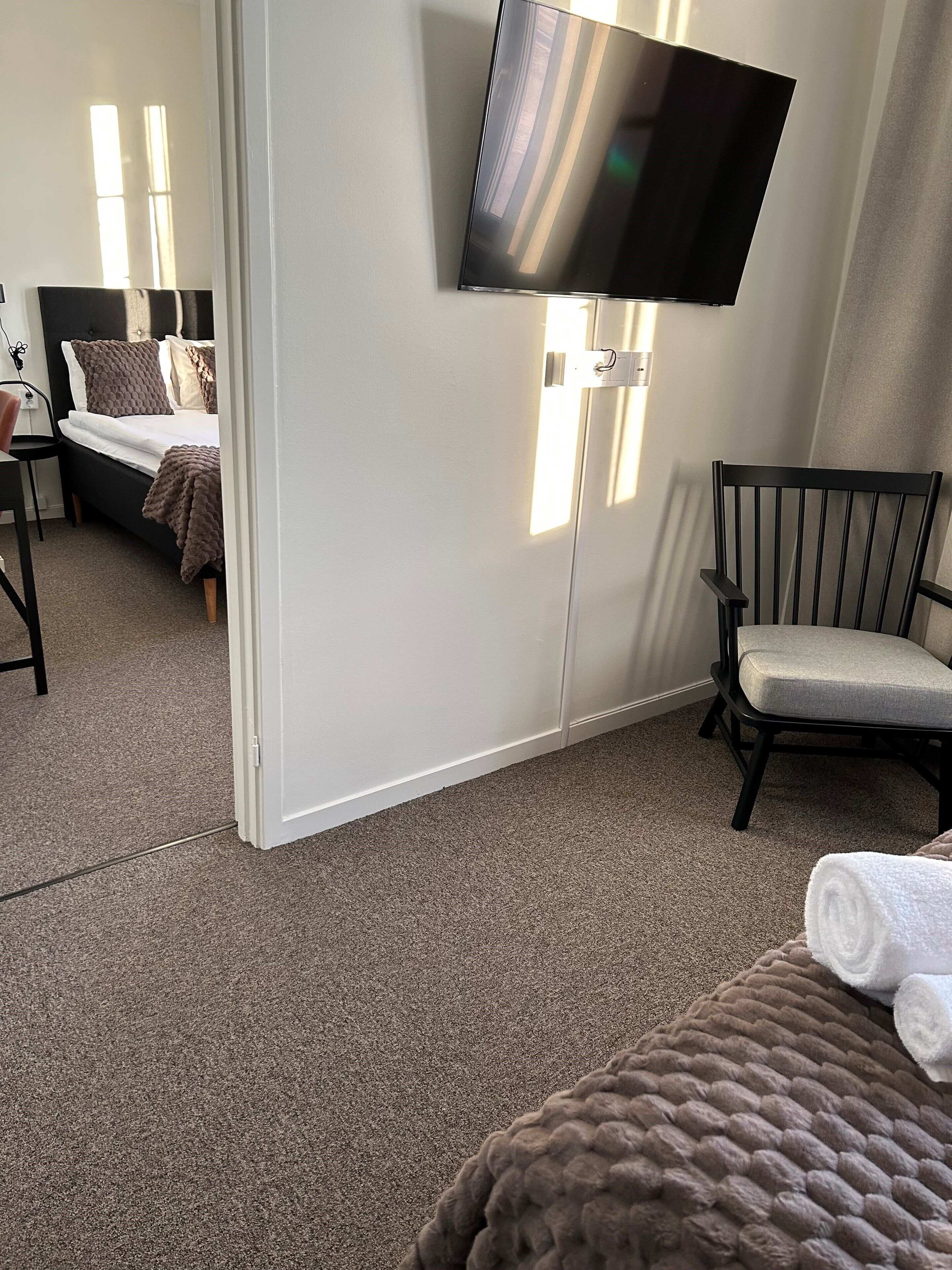 Quarto de hotel com cama, uma cadeira, televisão fixada na parede e uma mesa pequena.