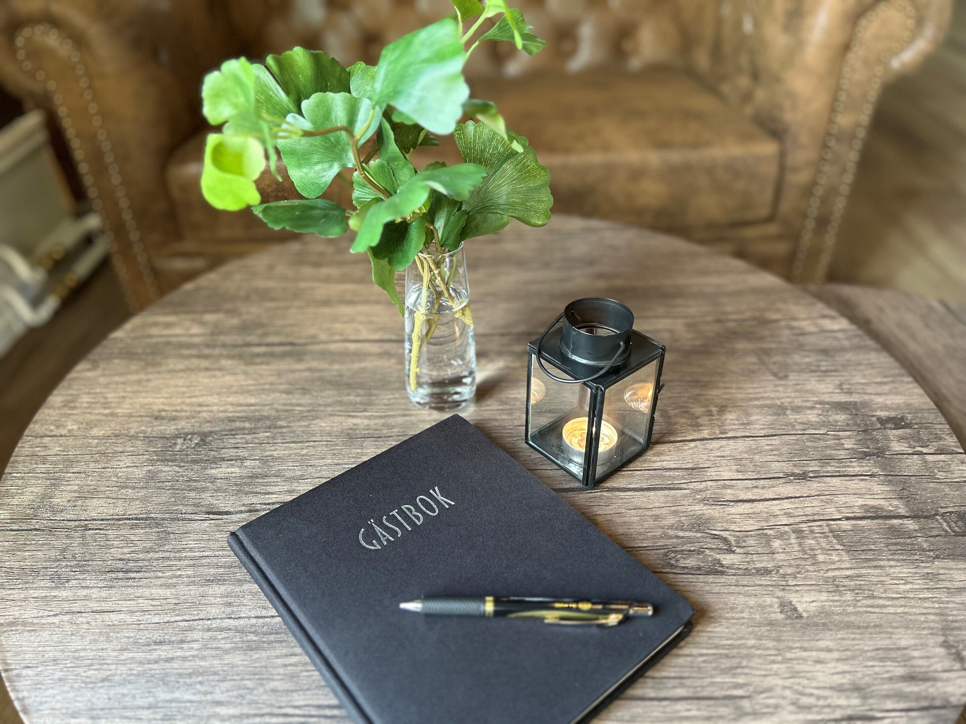 A wooden table with a black notebook labeled "GASTBOK," a pen, a small glass vase with green leaves, and a black lantern.