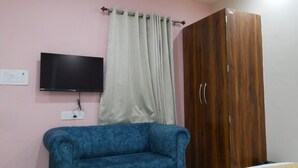 Quarto com sofá azul, guarda-roupa de madeira, televisão na parede e cortina.