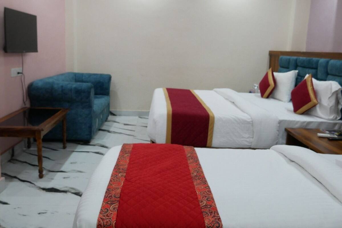 Habitación de hotel con dos camas, un sillón azul, una mesita de madera y un televisor de pantalla plana montado en la pared.