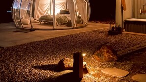 Une tente en forme de dôme transparent, avec un lit à l’intérieur, installée sur une surface de gravier, à proximité d’un petit abri.