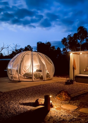 Une tente en forme de dôme transparent, avec un lit à l’intérieur, installée sur une surface de gravier, à proximité d’un petit abri.