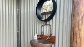 Une salle de bain avec un lavabo en pierre posé sur un tronc d’arbre, un miroir et un petit piédestal blanc.