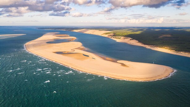 Vista aérea de un paisaje costero con playa arenosa, aguas azul cristalino y vegetación distante.