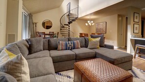 A spacious living room with a large sectional sofa, a wooden ottoman, a spiral staircase, and a dining area with a round table and chairs. - SkyRun Copper Mountain Resort Vacation Lodging (Copper Mountain)
